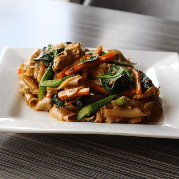 A white rectangular plate holds a serving of Thai noodles with chicken, carrots, and green vegetables.