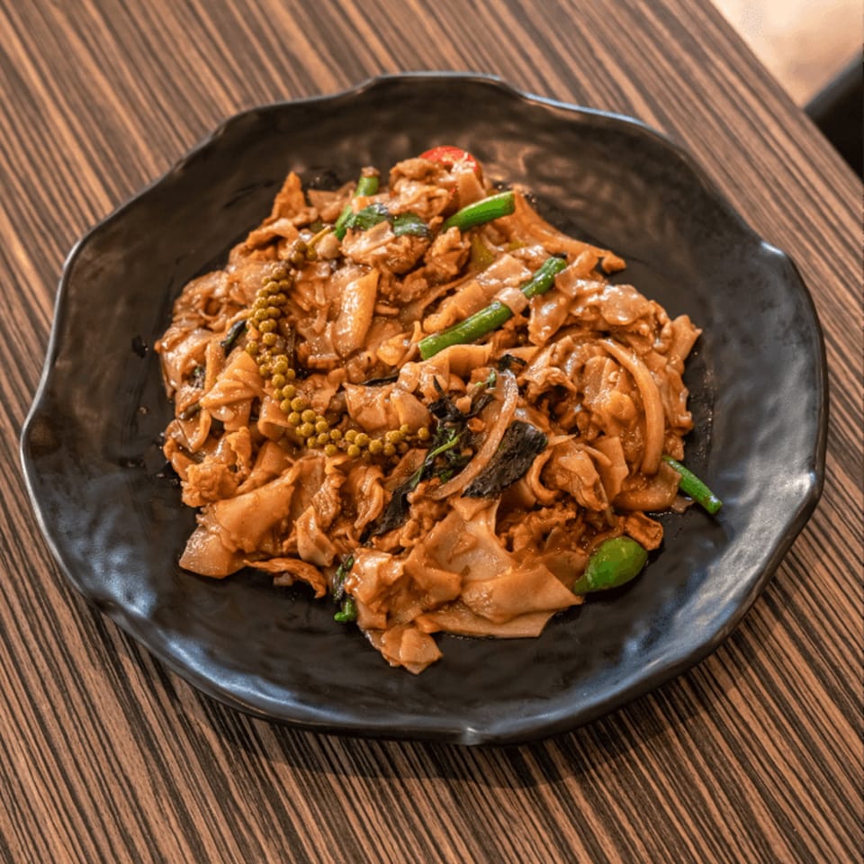 Spicy Thai drunken noodles with wide rice noodles, green peppercorns, and vegetables on a black plate.